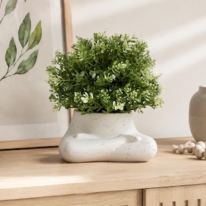 May include: A white ceramic planter with a speckled finish shaped like a figure sitting in a cross-legged position. The planter holds a dense, rounded artificial green plant with small leaves, creating a compact and balanced decorative display.