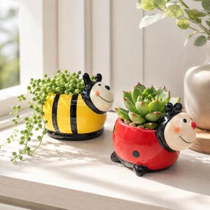 May include: Two ceramic animal-shaped planters. One is a yellow and black striped bee holding a trailing string of pearls plant. The other is a red ladybug with black spots holding a green succulent. Both have white faces with painted smiles.