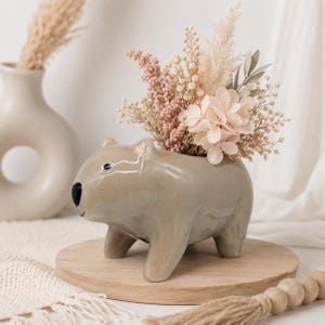 May include: A glossy, light brown ceramic planter shaped like a wombat, featuring a black nose and small ears. The planter is filled with a decorative arrangement of dried flowers in shades of cream, beige, and soft pink, including pampas grass.