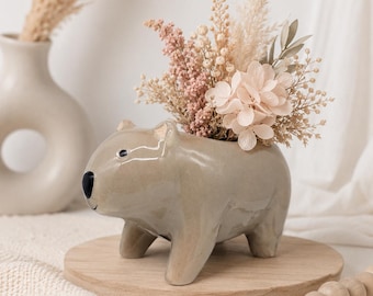 Op de afbeelding: Een glanzende, lichtbruine keramische plantenbak in de vorm van een wombat, voorzien van een zwarte neus en kleine oren. De plantenbak is gevuld met een decoratief arrangement van gedroogde bloemen in crème, beige en zachtroze tinten, inclusief pampasgras.