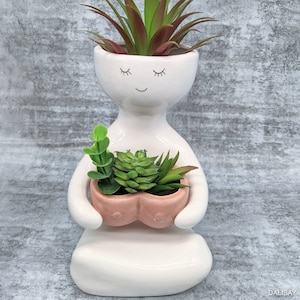 Könnte beinhalten: Weißer Keramik-Pflanztopf in Form einer Person mit geschlossenen Augen und einem Lächeln, der einen rosafarbenen herzförmigen Pflanztopf mit grünen Sukkulenten hält.