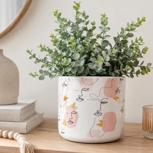 Puede incluir: Una maceta cilíndrica de cerámica blanca con un patrón de líneas negras de rostros abstractos, acentuado con formas en rosa suave y amarillo mostaza. La maceta contiene una planta de eucalipto verde con pequeñas hojas redondas.