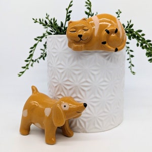 以下が含まれることがあります： セラミック製の猫と犬の置物セット。猫はオレンジと白で、黒い目と口を閉じています。犬は黄色で、白い斑点と黒い目があります。両方の置物は、幾何学模様の白いプランターの上に座っています。