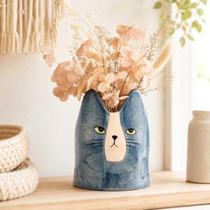Könnte beinhalten: Eine blaue Keramikvase in Form eines Katzenkopfes mit weißem Gesichtsfleck, gelben Augen und schwarzen Schnurrhaaren. Die Vase ist mit einem Strauß aus getrockneten beigen Blumen und Blattwerk gefüllt, mit zarten Blütenblättern und dünnen Stielen.