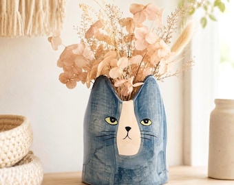 May include: A blue ceramic vase shaped like a cat head with a white face patch, yellow eyes, and black whiskers. The vase is filled with a bouquet of dried beige flowers and foliage, featuring delicate petals and thin stems extending upwards.