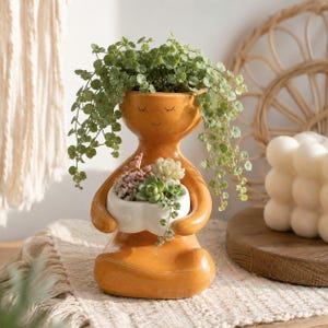 May include: A glossy mustard-yellow ceramic planter shaped like a person sitting in a cross-legged position. The figure holds a small white heart-shaped bowl containing various succulents, while a trailing green plant spills from the head area.