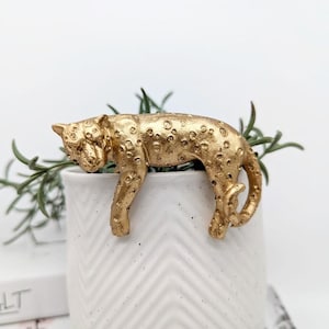 May include: A gold-painted figurine of a leopard sleeping on a white planter with a chevron pattern. The planter has a green plant growing out of it.