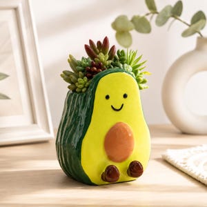 May include: A ceramic avocado-shaped planter with a glossy finish, featuring a bright yellow centre, dark green textured sides, a brown pit, and two small brown feet. The planter contains a variety of small green and reddish succulent plants.