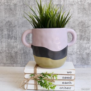 Puede incluir: Una pila de libros blancos con el texto "we need each other" impreso en el libro superior. Los libros están atados con cuerda y una pequeña planta verde se coloca encima de los libros. Una maceta de cerámica de color púrpura claro con rayas onduladas negras y doradas y dos asas está encima de los libros. Una planta verde está creciendo en la maceta.