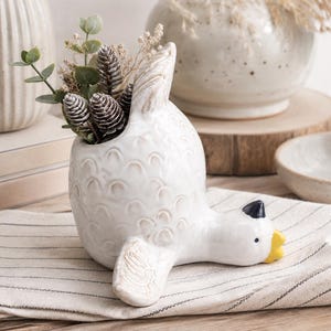 May include: A white ceramic goose-shaped planter with a textured feather pattern on its body. The goose has a yellow beak and a black tip, with a hollow opening on its back containing small pinecones and green eucalyptus sprigs.