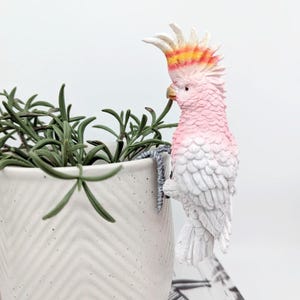 May include: A pink and white cockatoo figurine perched on the edge of a white ceramic planter with a chevron pattern. The cockatoo has a yellow and orange crest.