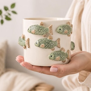 May include: A cream-colored ceramic planter adorned with six green and tan fish. The fish are arranged around the planter's exterior. The planter is cylindrical and appears to be held by a person.