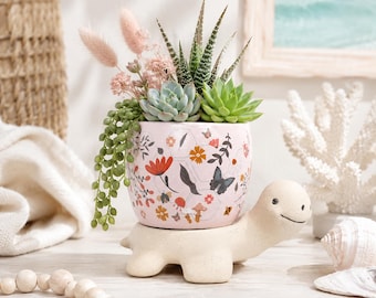 May include: A light pink ceramic planter shaped like a turtle, featuring a colourful floral and butterfly pattern. The planter holds a variety of succulents and dried pink grasses, with a trailing green plant cascading down the side of the pot.