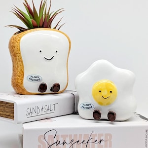 May include: Two ceramic planters: a slice of toast with a plant and a fried egg. Both have smiling faces and the text "Funky Friends". The toast planter is brown and white, and the egg planter is white and yellow. They are decorative items.