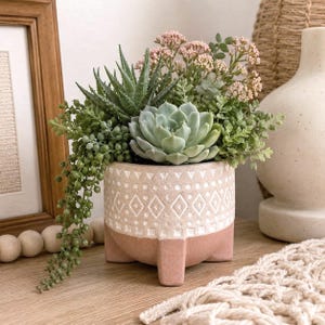 May include: A round ceramic planter with three short legs, featuring a two-tone design with a white upper section displaying a geometric diamond pattern and a solid pink lower section, filled with an arrangement of various green artificial succulents.