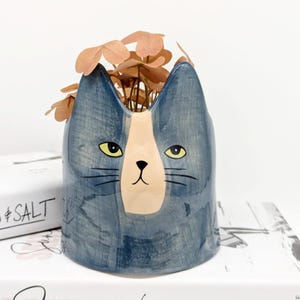 May include: A blue ceramic cat planter with a white and peach face. The planter has a glazed finish and is ideal for small plants.