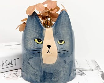 May include: A blue ceramic cat planter with a white and peach face. The planter has a glazed finish and is perfect for small plants.