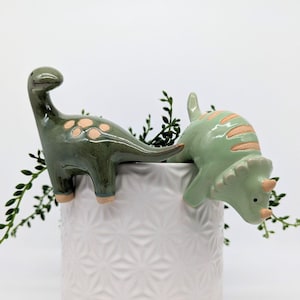 May include: Two ceramic dinosaur figurines, a green triceratops and a green brachiosaurus, are perched on the edge of a white planter with a geometric pattern. The planter is filled with green foliage.