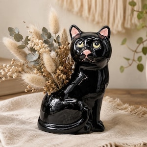 May include: A glossy black ceramic cat figurine shaped as a vase, featuring large yellow eyes and a pink nose. The cat is filled with a bouquet of dried beige bunny tail grass and small green eucalyptus leaves, creating a decorative floral display.