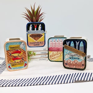 May include: Four ceramic sardine can-shaped planters with colourful designs. One planter has the text "TINNED FISH" and "SUCH A GREAT CATCH". Another says "SARDINES IN BRINE" and "PARTNERS". The third says "plenty MORE FISH". The last says "Together WE'RE BETTER".