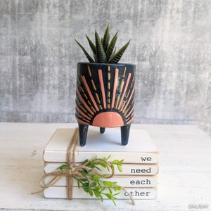 May include: A black and pink ceramic planter with a sun design and three legs. The planter is filled with a green succulent plant. The planter is sitting on top of a stack of white books with the text "we need each other" written on them.