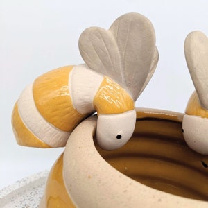 May include: A ceramic planter with a honey-coloured interior and exterior, featuring two stylised bees. The bees have yellow and white striped bodies and cream-coloured wings, adding a whimsical touch to the decorative piece.
