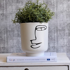 May include: White ceramic planter with a black line drawing of a face. The planter is filled with green leafy plants.