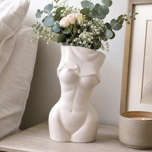May include: A cream-coloured ceramic vase shaped like a human torso with a speckled finish. The vase holds a bouquet of green eucalyptus leaves, white baby's breath, and small pale pink roses, creating a decorative floral arrangement piece.