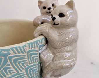 Puede incluir: Dos figuritas de cerámica de gato con glaseado blanco y ojos negros están posadas en el borde de una maceta de cerámica con estampado azul y blanco.