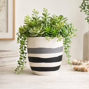 May include: A speckled off-white ceramic planter with horizontal gray and black painted stripes, filled with a variety of green artificial succulents and trailing foliage that spills over the rounded edges of the container.