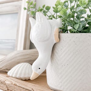 May include: A white ceramic goose figurine designed to hang over the edge of a planter. The goose has a smooth finish, a small black eye, and a light tan beak. It is positioned on the rim of a speckled, off-white cylindrical plant pot.