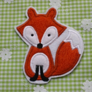Könnte beinhalten: Ein Fuchsapplikation aus Filz in Orange, Weiß und Schwarz. Der Fuchs hat große weiße Augen, eine schwarze Nase und einen flauschigen weißen Schwanz. Die Applikation befindet sich auf einem grün-weiß karierten Hintergrund mit weißen Blumenakzenten.
