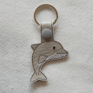 Könnte beinhalten: Ein delfinförmiger Schlüsselanhänger aus strukturiertem hellgrauem Material mit dunkelgrauen Nähten an den Rändern. Das Design zeigt ein kleines schwarzes Auge und silberfarbene Stickereidetails am Flossenbereich, befestigt mit einem grauen Druckknopf und einem Metallring.