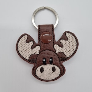 Könnte beinhalten: Brauner Leder-Elch-Schlüsselanhänger mit einem weißen gestickten Gesicht und Geweih. Der Schlüsselanhänger hat einen silbernen Metallring.