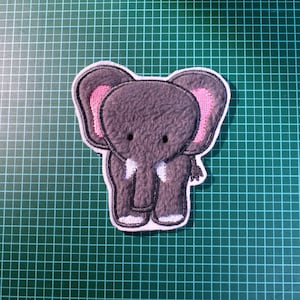 Könnte beinhalten: Ein grauer Filz-Elefanten-Applik mit rosa Ohren und weißen Stoßzähnen. Der Elefant steht auf einem grünen Hintergrund.