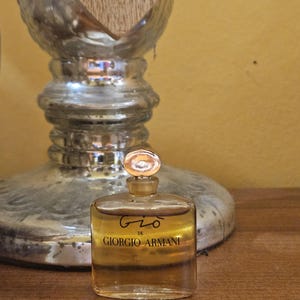 Pode incluir: Um pequeno frasco retangular de perfume "Gio" de Giorgio Armani com tampa e rótulo dourados. O frasco contém um líquido âmbar claro. O texto "Gio de Giorgio Armani" está impresso na frente. O frasco está sobre uma superfície de madeira.