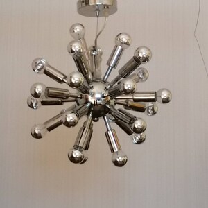 Könnte beinhalten: Eine verchromte Sputnik-Stil-Deckenleuchte mit mehreren Glühbirnen. Die Leuchte hat eine zentrale Kugel mit radialen Armen, die jeweils eine Glühbirne halten. Die Glühbirnen sind klar, und das Metall hat eine glänzende, reflektierende Oberfläche. Die Deckenleuchte ist von der Decke abgehängt.