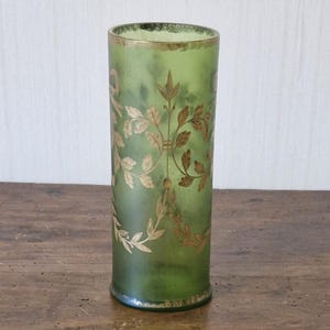 May include: A cylindrical green glass vase with gold leaf designs. The vase has a slightly textured surface and a gold rim. It stands on a wooden surface. The design features floral and leaf patterns.
