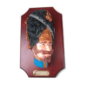 Puede incluir: Escultura mural tridimensional que representa la cabeza de un granadero de la Guardia Imperial. La escultura presenta un sombrero de piel negro, un casco color cobre y un bigote marrón rojizo. Montada sobre una placa de madera oscura.