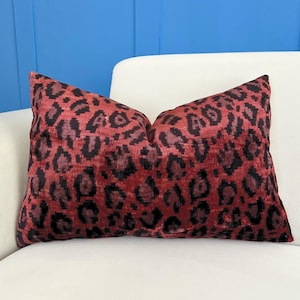 May include: A rectangular decorative pillow with a red and black leopard print pattern. The pillow is placed on a white sofa. The background features a blue wall.