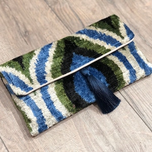Puede incluir: Un bolso de mano rectangular con textura de terciopelo y un llamativo patrón ikat en tonos verde oscuro, azul real, negro y blanco roto. La solapa cuenta con una borla azul oscuro a juego y un ribete de color claro en los bordes.