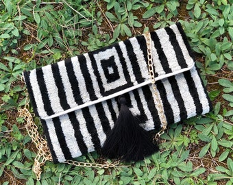 Puede incluir: Clutch de rayas blancas y negras con una correa de cadena dorada y un borla negra. La bolsa tiene un patrón geométrico en la parte delantera.