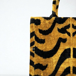 May include: A rectangular tote bag with a gold and black patterned design. The bag features a repeating pattern of curved black lines on a gold background. The handles are also gold and black.
