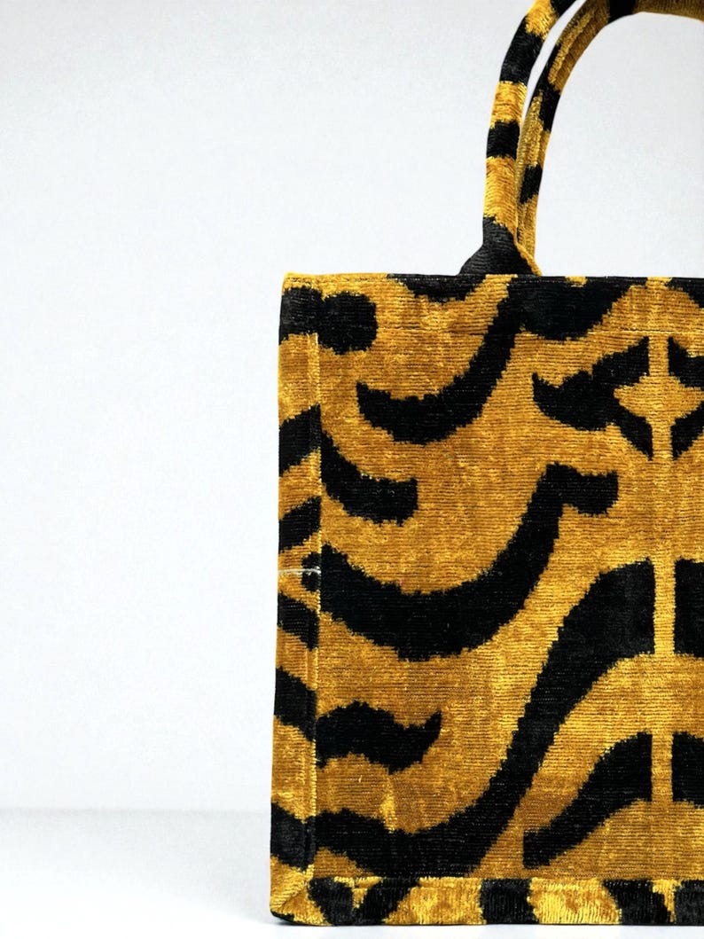 May include: A rectangular tote bag with a gold and black patterned design. The bag features a repeating pattern of curved black lines on a gold background. The handles are also gold and black.