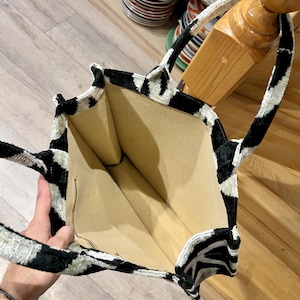 May include: A black and white patterned tote bag with a zebra-like design. The bag has a beige interior and a zipper closure. The handles are also black and white. The bag is open, revealing the interior.