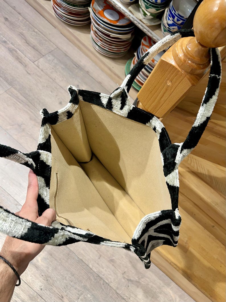 May include: A black and white patterned tote bag with a zebra-like design. The bag has a beige interior and a zipper closure. The handles are also black and white. The bag is open, revealing the interior.