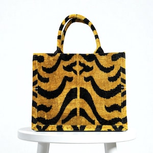 May include: A rectangular tote bag with a tiger stripe pattern. The bag features a mustard yellow background with black curved stripes. The handles are also yellow with black stripes. The bag is sitting on a white surface.