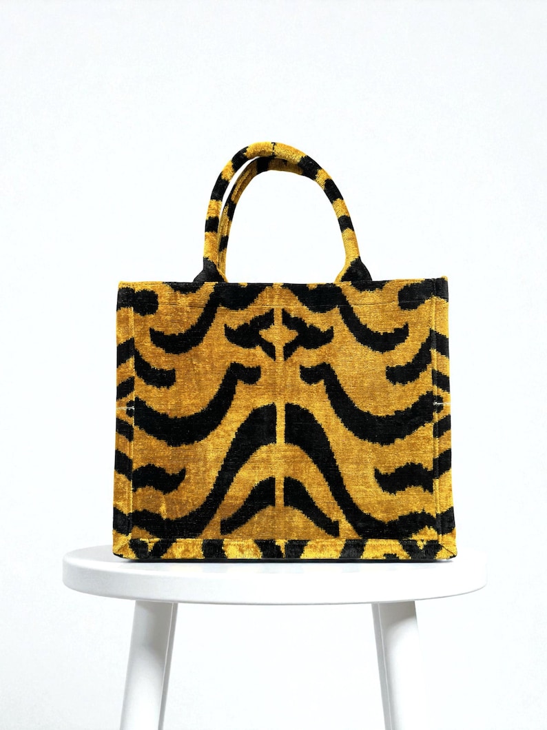 May include: A rectangular tote bag with a tiger stripe pattern. The bag features a mustard yellow background with black curved stripes. The handles are also yellow with black stripes. The bag is sitting on a white surface.