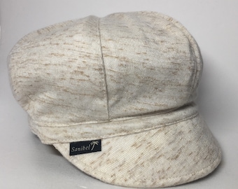 May include: A beige knitted newsboy cap with a brown and white speckled pattern. The cap has a black label with the word "Sanibel" and a palm tree logo.