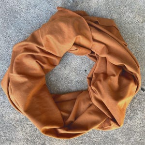 May include: A circular, rust-coloured infinity scarf made of soft fabric. The scarf is draped in a circular shape, showcasing its texture and colour. The warm, earthy tone suggests a cosy accessory for cooler weather.